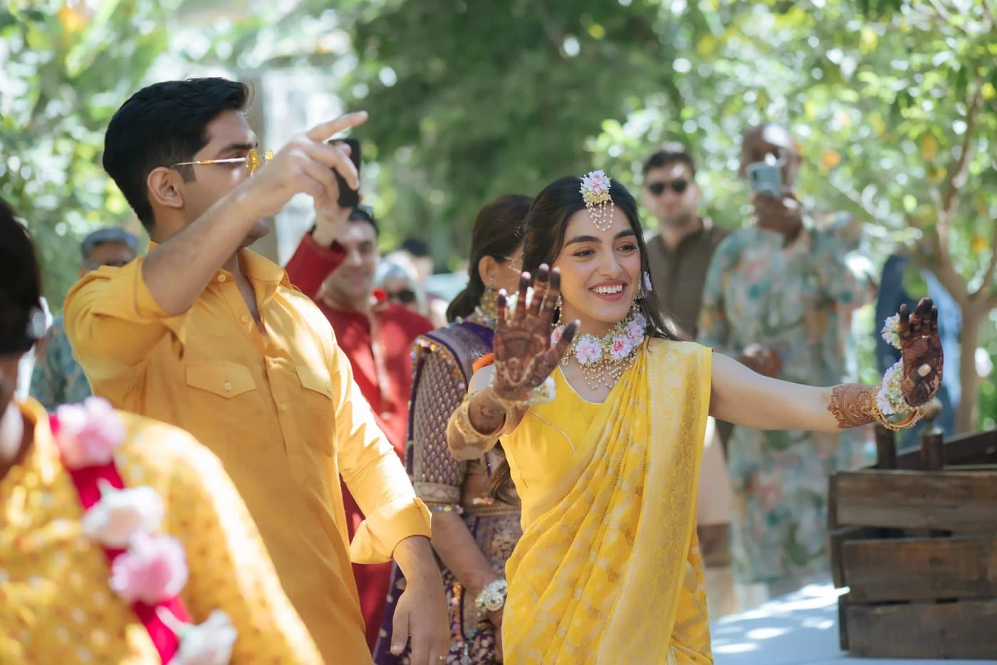 A couple in yellow dancing along a tree-lined path during a mehndi procession.
