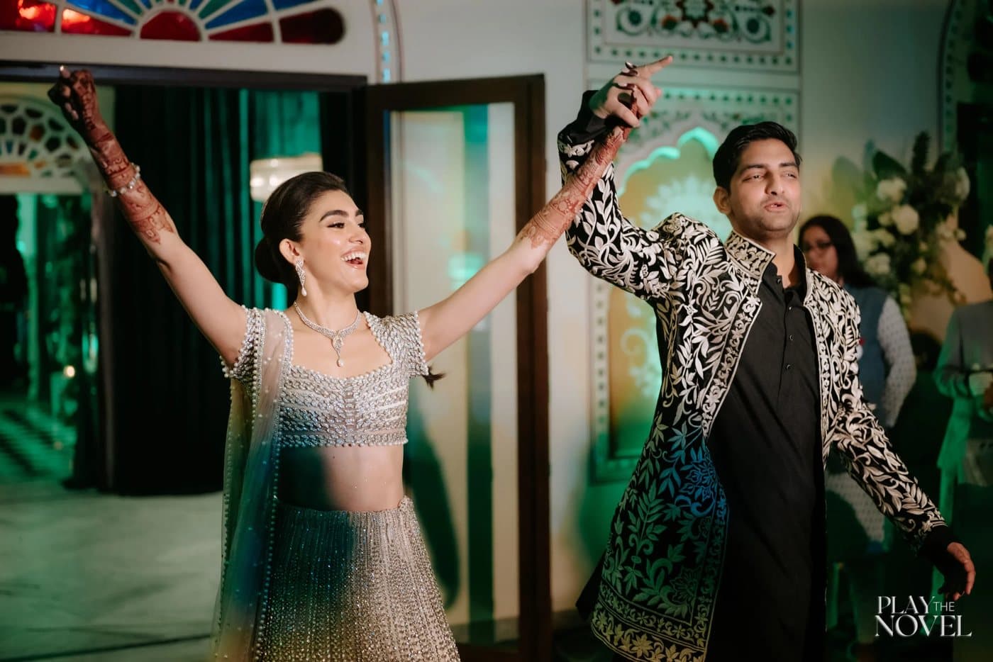 A bride in a silver lehenga throwing her arms open mid-celebration.
